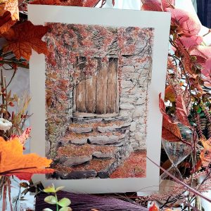 Aquarelle originale présentée sur chevalet représentant un bâtiment en pierre avec une vieille porte en bois et des feuilles mortes au sol @Ysae illustrations