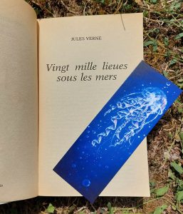 Photo du livre "20 000 lieues sous les mers" avec un mini marque-page illustré représentant une méduse iridescente dans le bleu profond de l'océan @ysae illustrations