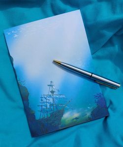 Bloc-note illustré représentant un galion noyé au fond de l'océan, les voiles déchirées. Il est présenté avec un stylo sur une serviette de plage @ysae illustrations