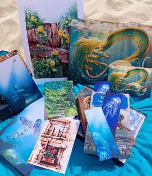 Photo du pack de dessins d'été avec des affiches, des cartes postales, un bloc-notes et des stickers présentés avec des livres sur la plages. Ce sont des dessins numériques, des peintures gouaches et aquarelles représentant la nature, la mer et la faune sous-marine @ysae illustrations