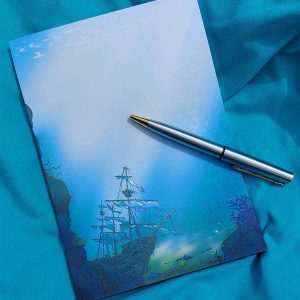 Bloc-note illustré représentant un galion noyé au fond de l'océan, les voiles déchirées. Il est présenté avec un stylo sur une serviette de plage @ysae illustrations