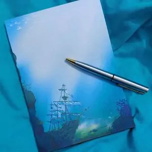 Bloc-note illustré représentant un galion noyé au fond de l'océan, les voiles déchirées. Il est présenté avec un stylo sur une serviette de plage @ysae illustrations